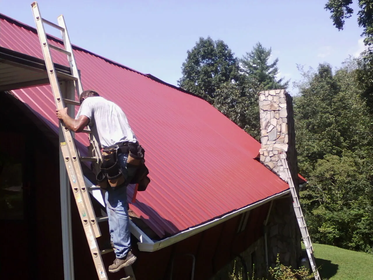 Roofer on a ladder beside a red metal roof and stone chimney — Soffit & Fascia Repair by Roofing Brothers in Shannon